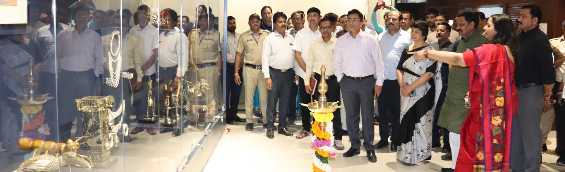 आदिवासी पितळी कलावस्तू, मुखवटे व दागिने ठेवलेल्या काचेच्या कपाटासमोर उभे सुमारे पंचवीस अधिकारी; शेजारी झेंडूच्या माळांनी सजवलेली प्रज्वलित समई आणि लाल साडीतील महिला कपाटाकडे निर्देश करताना