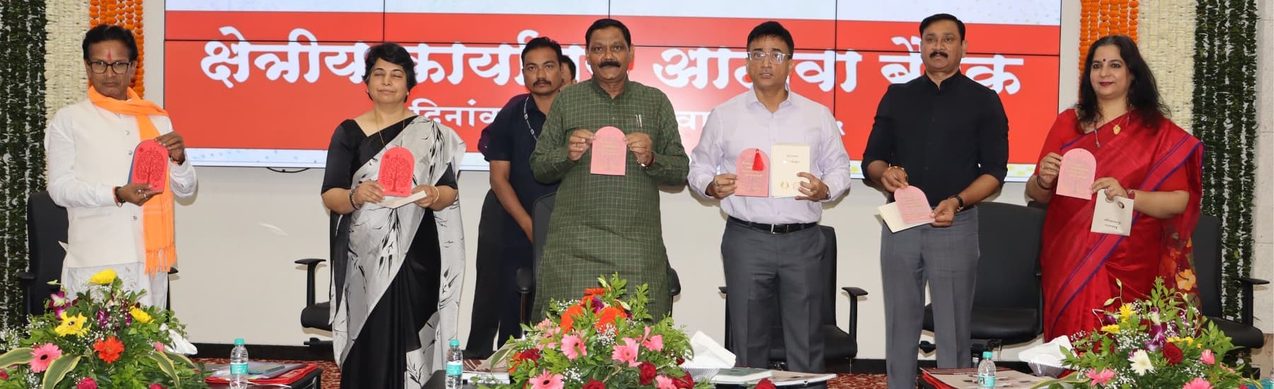 'क्षेत्रीय कार्यालय आढावा बैठक' असे लिहिलेल्या पडद्यासमोर व्यासपीठावर सात मान्यवर कमानीच्या आकाराची गुलाबी पुस्तिका उंचावून प्रकाशन करताना