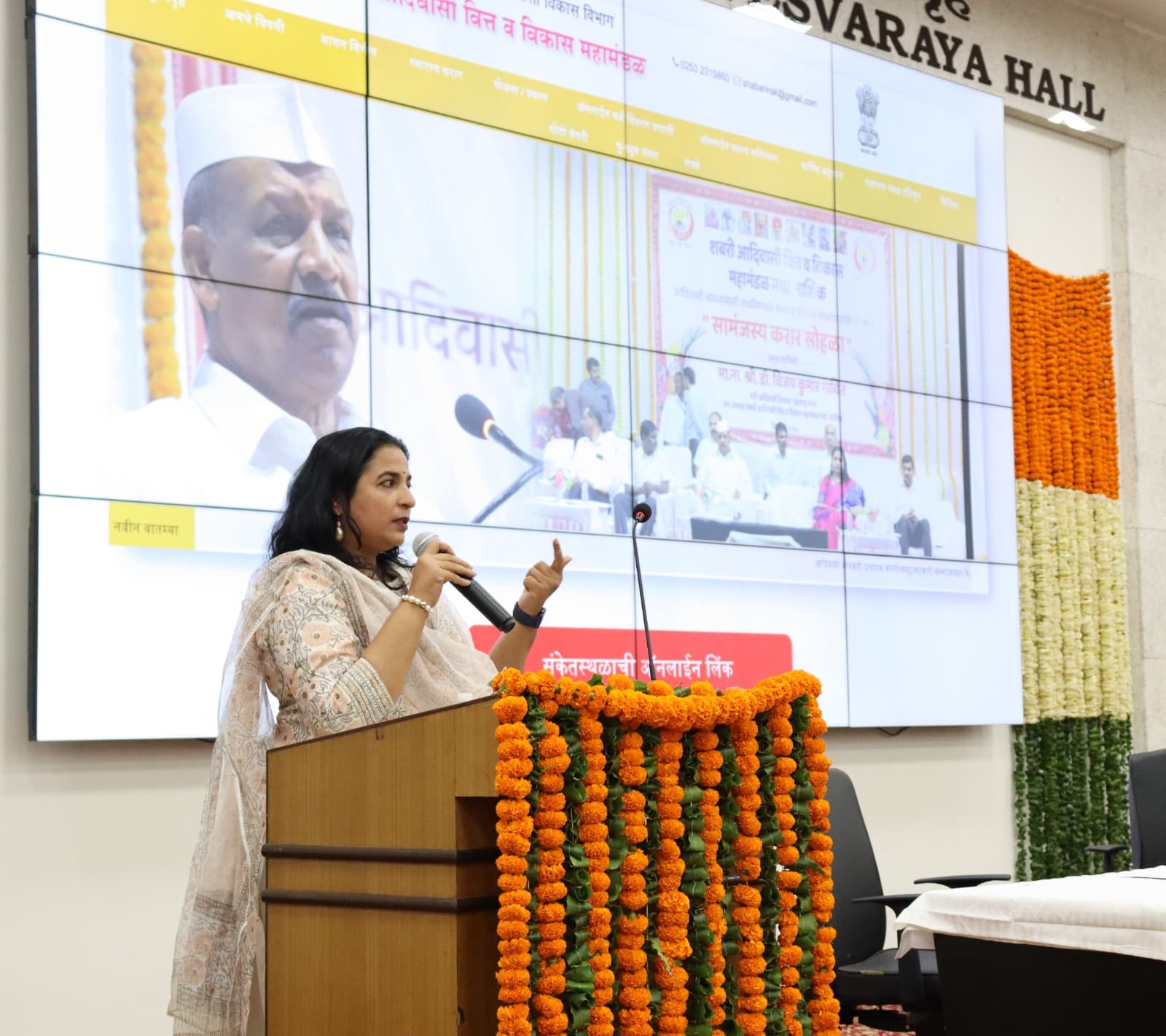 व्यासपीठावरून बोलणाऱ्या महिला अधिकारी; मागील पडद्यावर लोकार्पण होत असलेल्या महामंडळाच्या संकेतस्थळाचे मुखपृष्ठ — मराठी मेनू, दूरध्वनी क्रमांक व ई-मेल दिसत आहे