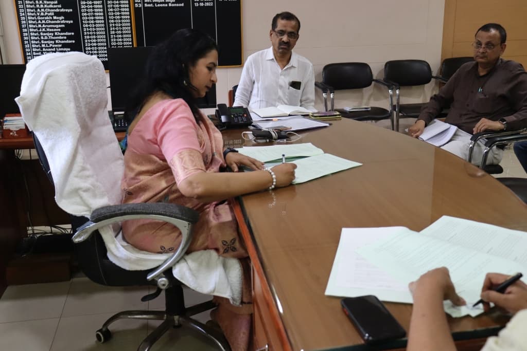लाकडी टेबलावर हिरव्या पुठ्ठ्याचे दस्तऐवज स्वाक्षरी करणाऱ्या गुलाबी साडीतील महिला अधिकारी; समोर दोन पुरुष आणि मागील भिंतीवर पूर्वीच्या पदाधिकाऱ्यांचा नामफलक