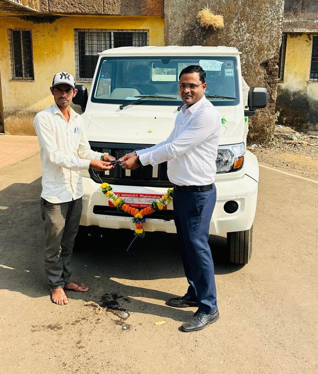 जुनाट पिवळ्या इमारतीसमोर हार घातलेल्या नव्या बोलेरो पिकअपच्या बोनेटवरून टोपी घातलेल्या लाभार्थ्याला चावी देणारे अधिकारी