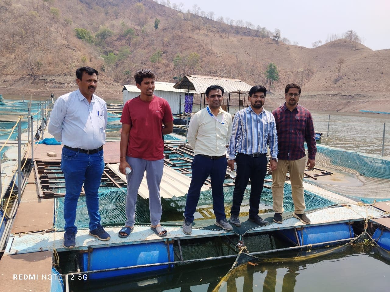 जलाशयातील निळ्या पिंपांवरच्या तरंगत्या मत्स्यपिंजरा फळीवर उभे पाच जण; मागे उघडे डोंगर व तरंगती पत्र्याची खोली
