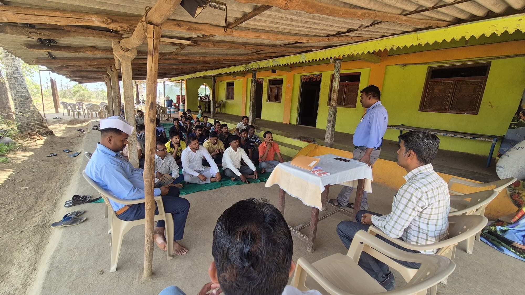 रंगवलेल्या गावातील घराच्या ओसरीवर हिरव्या सतरंजीवर बसलेले सुमारे पंचवीस पुरुष आणि त्यांना संबोधित करणारा निळ्या शर्टातील अधिकारी